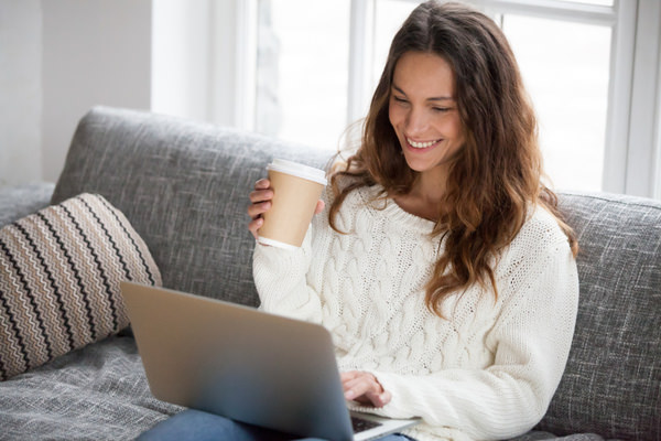 A woman sits on her couch with a cup of coffee while on her laptop