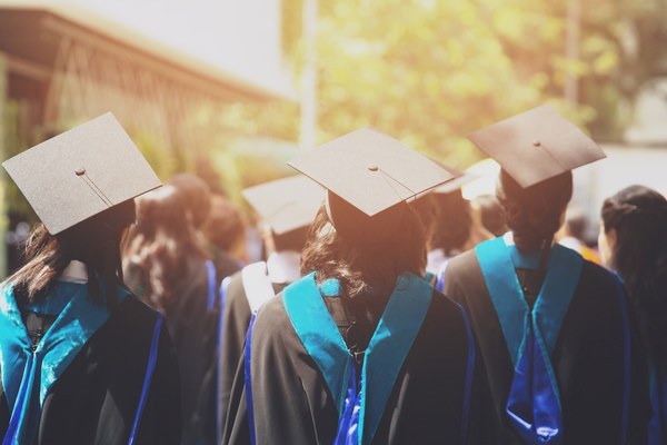 Three graduates stand with their backs to the camera. The are wearing robes and hats.