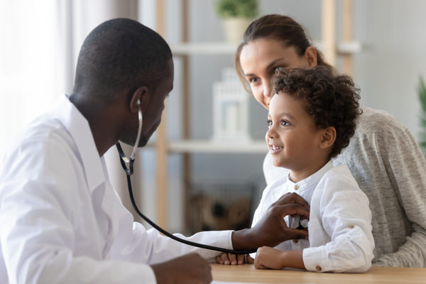 A doctor holds a stethescope to a young boy's chest. The boy is sitting on his mothers lap.