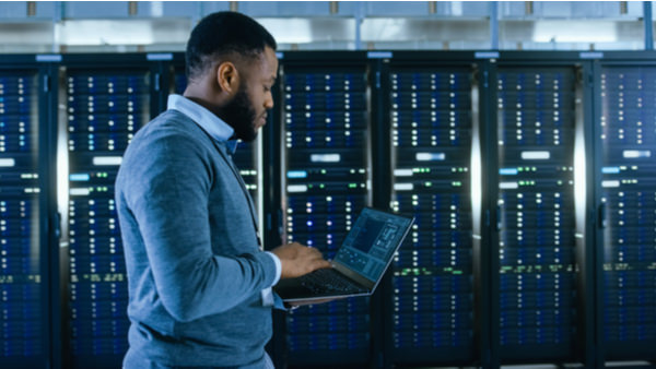 A man on his laptop stands in front of a wall of servers